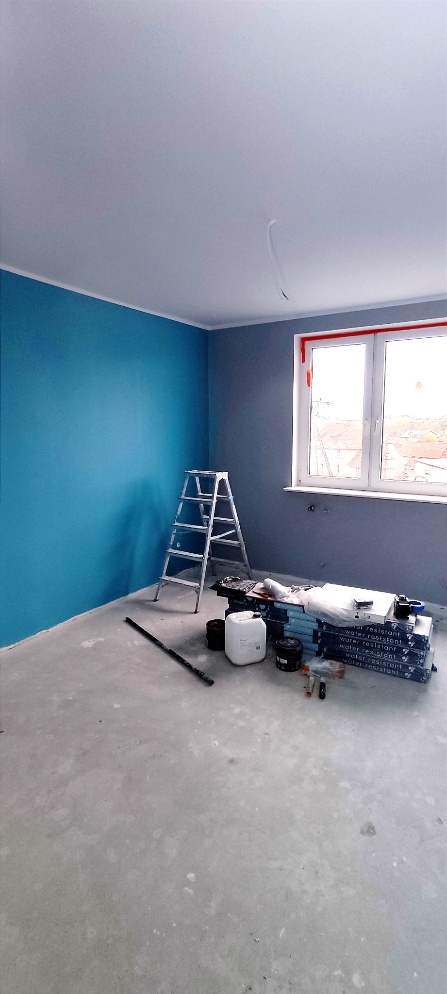 Świeżo pomalowane pomieszczenie z błękitną ścianą, szarą ścianą przy oknie, białą listwą sufitową, aluminiową drabiną, wałkiem, opakowaniami paneli podłogowych, kanistrem i puszkami farby.