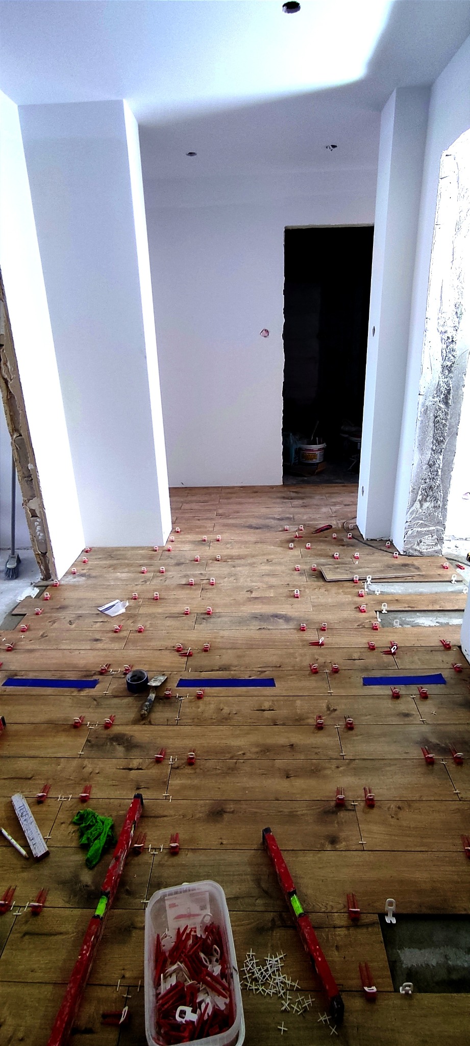 Układanie płytek imitujących drewno, widoczne poziomice, krzyżyki dystansowe, niebieska taśma maskująca, pomieszczenie w trakcie prac wykończeniowych.
