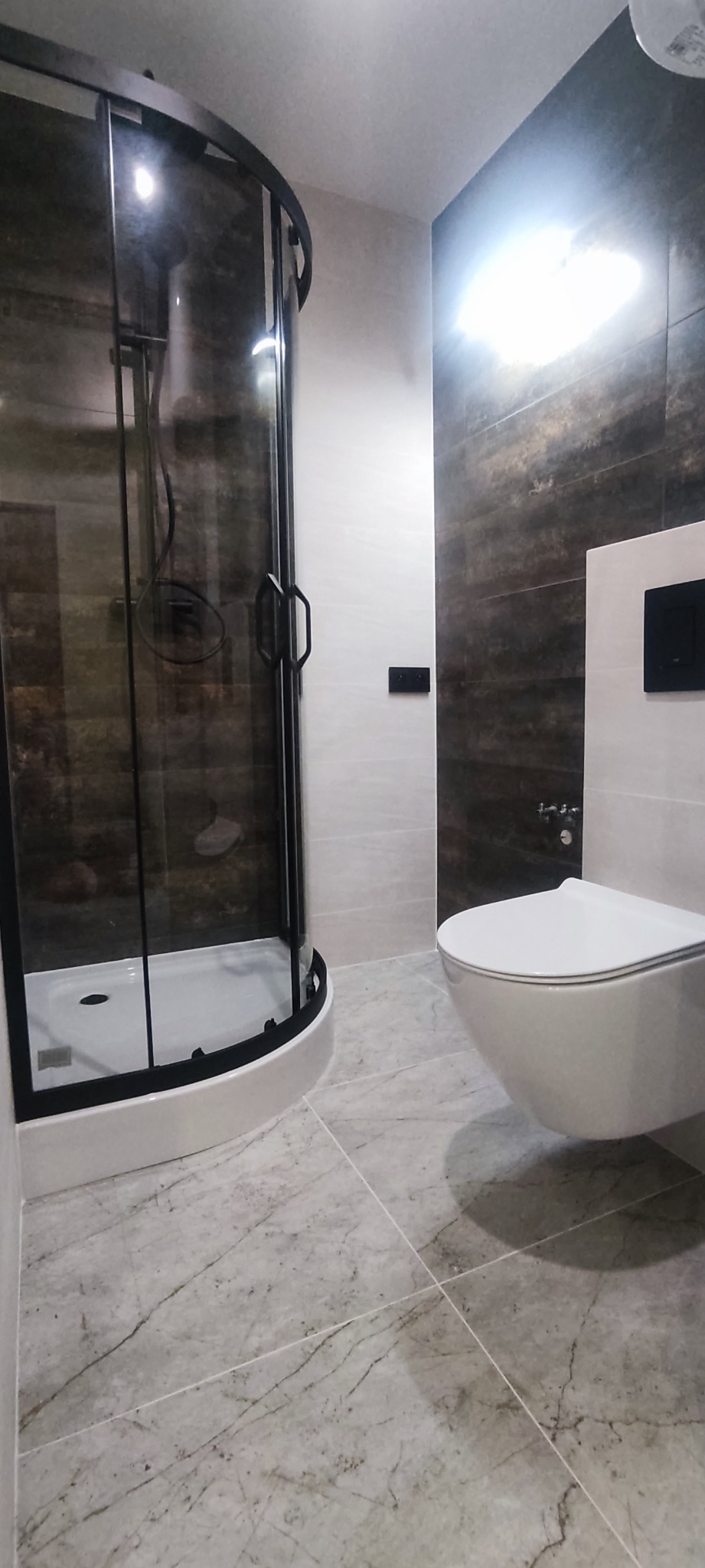 Nowoczesna łazienka z kabiną prysznicową typu walk-in z czarnymi profilami, podwieszaną toaletą i marmurową posadzką.
