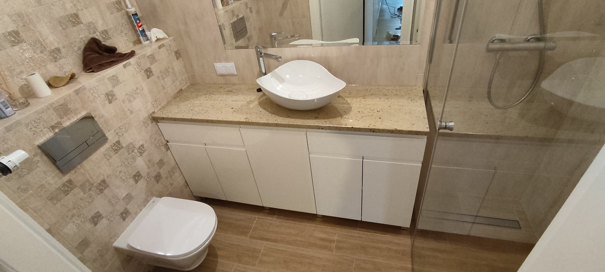 Nowoczesna łazienka z beżowymi płytkami, owalną białą umywalką na blacie z jasnego kamienia, białymi szafkami pod umywalką i kabiną prysznicową ze szklanymi drzwiami