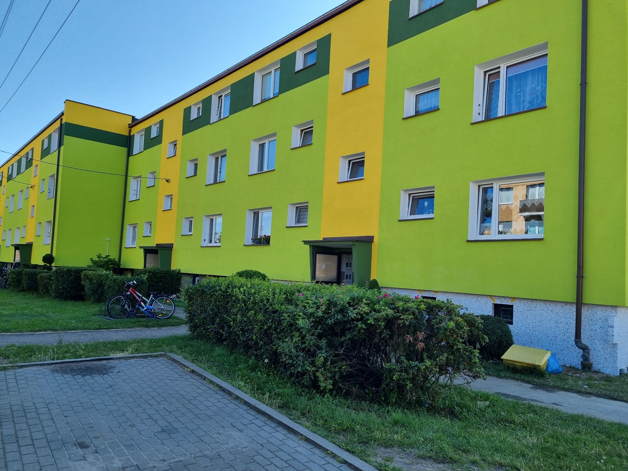 Wielopiętrowy budynek mieszkalny po renowacji elewacji, pomalowany w jaskrawe kolory: limonkową zieleń i żółty, z białymi ramami okiennymi, widok z poziomu chodnika z zaparkowanymi rowerami w tle.