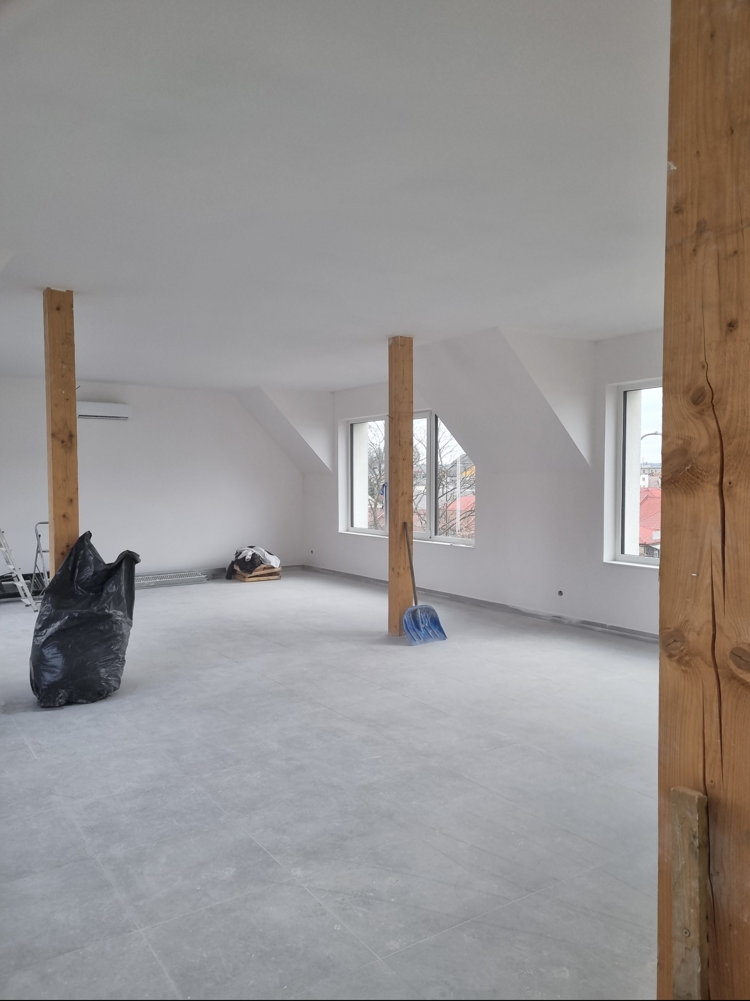 Poddasze po remoncie z drewnianymi belkami, świeżo pomalowanymi ścianami, szarymi płytkami podłogowymi, workiem na śmieci i niebieską łopatą.