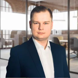 Portret mężczyzny w ciemnym garniturze i białej koszuli, na rozmytym tle nowoczesnego biura z przeszklonymi ścianami i widoczną salą konferencyjną.