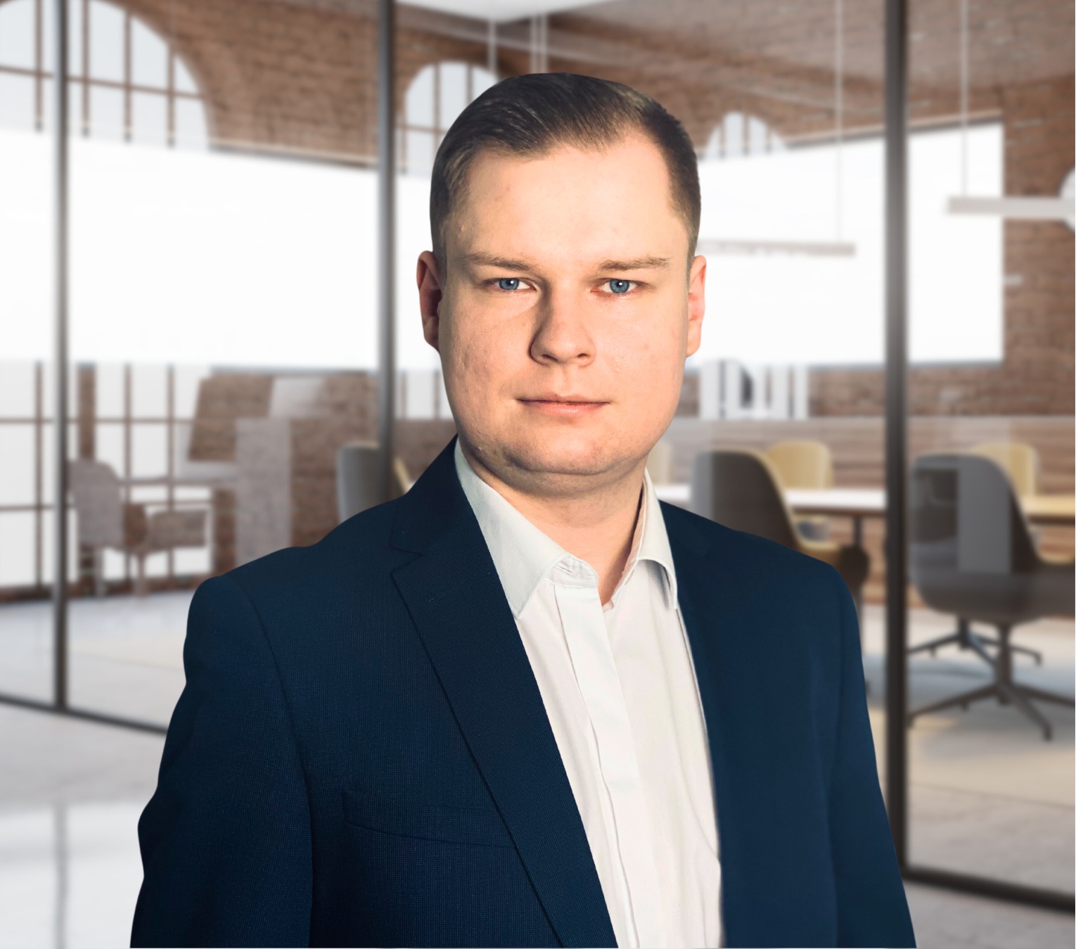 Portret mężczyzny w ciemnym garniturze i białej koszuli, na rozmytym tle nowoczesnego biura z przeszklonymi ścianami i widoczną salą konferencyjną.