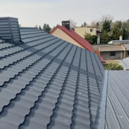 Nowy, grafitowy dach z blachodachówki modułowej z kominem wentylacyjnym, widok z góry na tle zabudowy jednorodzinnej.