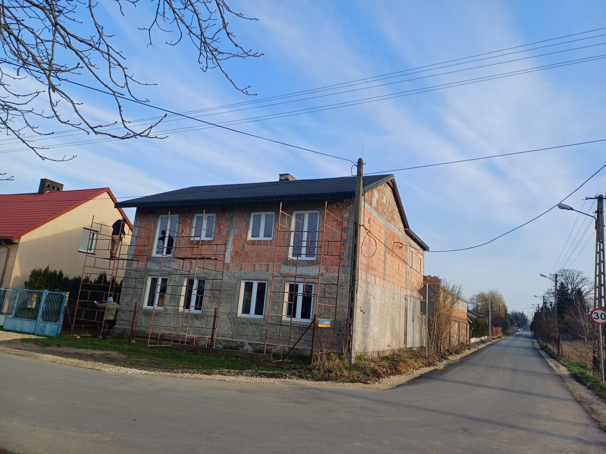Dwupiętrowy dom w trakcie budowy z nowym, czarnym dachem, otoczony rusztowaniami, widoczny z perspektywy ulicy.