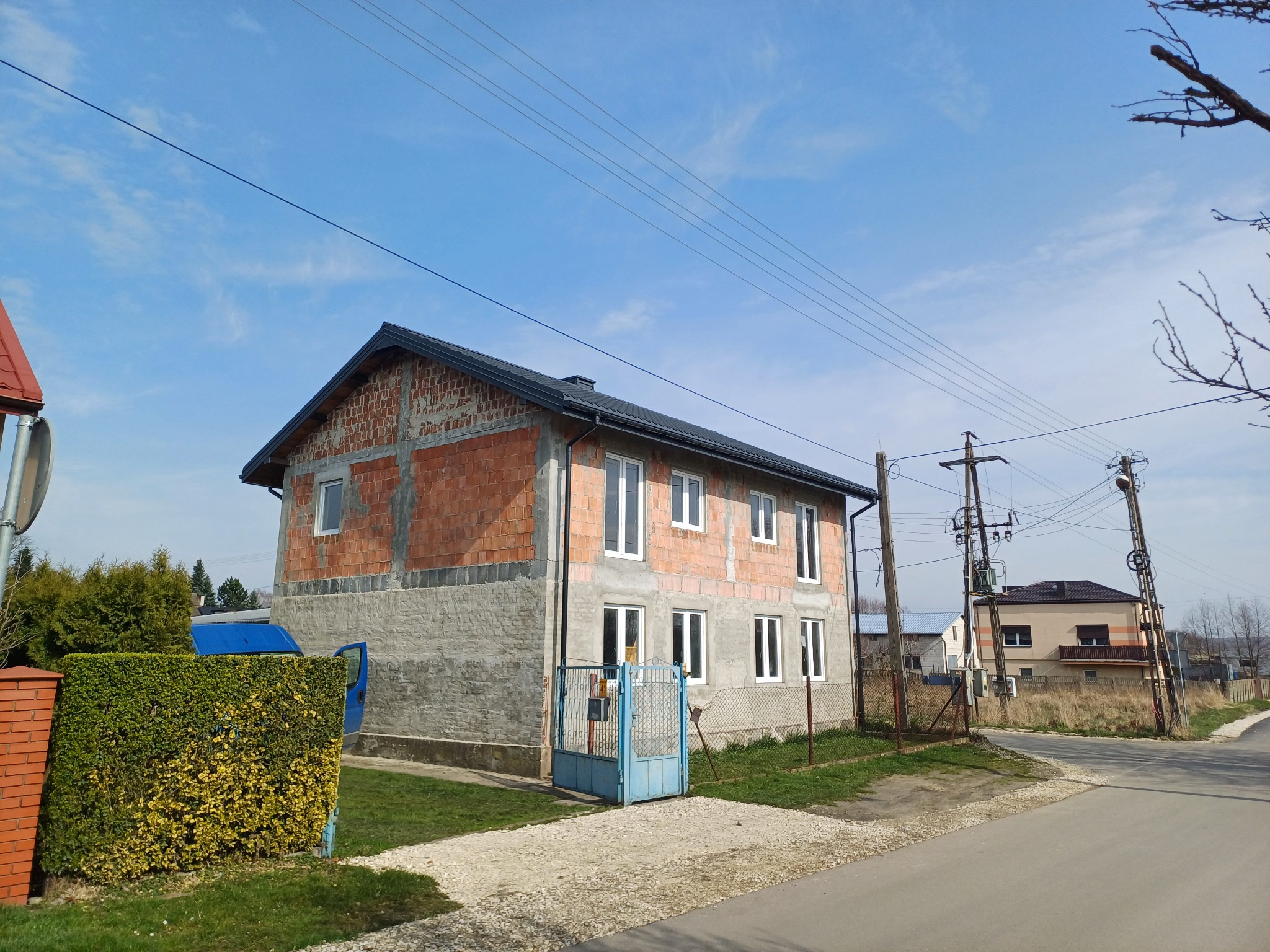 Dwukondygnacyjny dom w trakcie budowy z widoczną surową cegłą na górnej kondygnacji i szarym tynkiem na dole, z nowym, ciemnym dachem i białymi oknami, na tle błękitnego nieba.