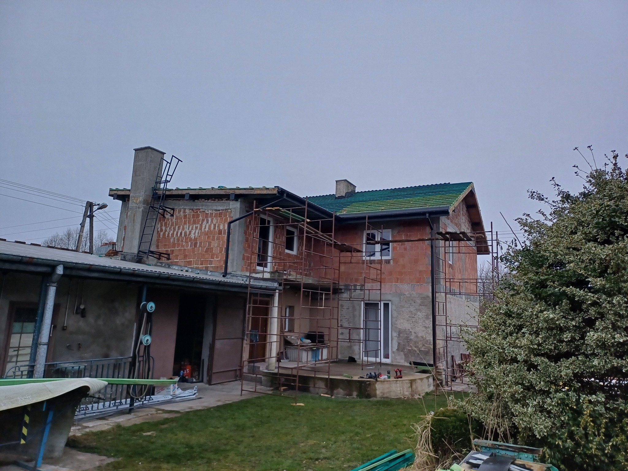 Dom jednorodzinny w trakcie prac dekarskich, widoczne rusztowania, częściowo położone zielone pokrycie, ceglany komin z drabinką, oraz przygotowane narzędzia na trawniku.
