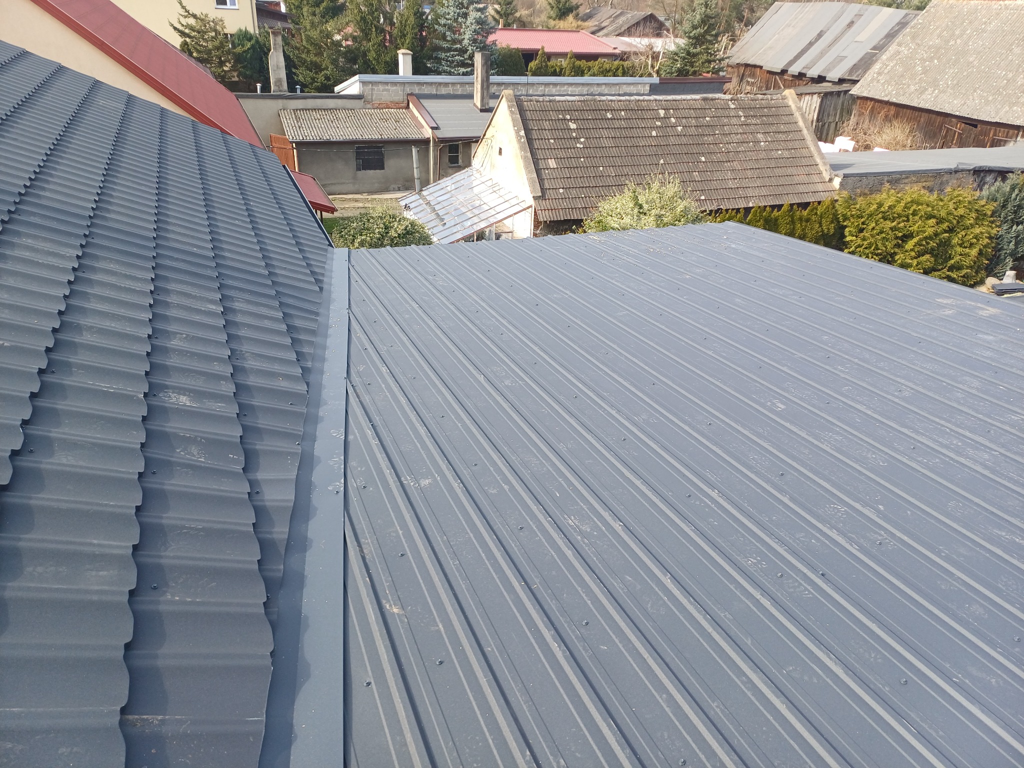 Szary, metalowy dach z widocznym łączeniem blach, w tle zabudowa jednorodzinna i drzewa.