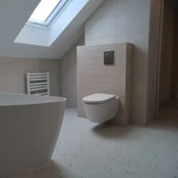 Nowoczesna łazienka na poddaszu z oknem dachowym, wolnostojącą wanną, podwieszaną toaletą i płytkami imitującymi lastryko na podłodze.