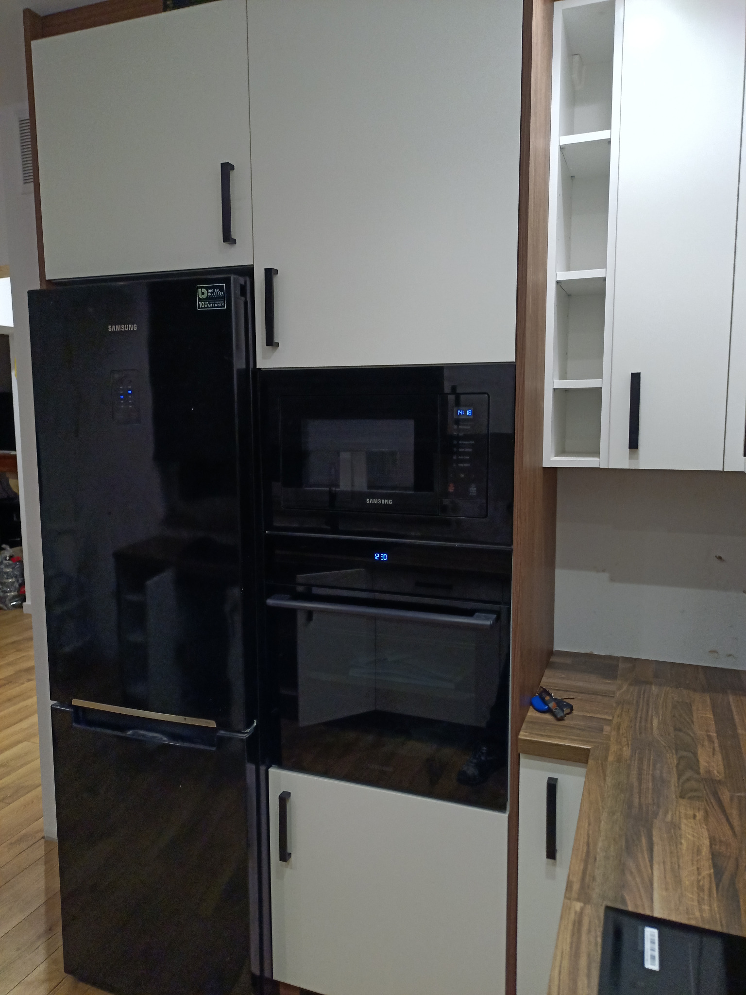 Nowoczesna kuchnia z zabudową meblową w odcieniach bieli i drewna, z czarnym sprzętem AGD marki Samsung: lodówką, mikrofalówką i piekarnikiem.