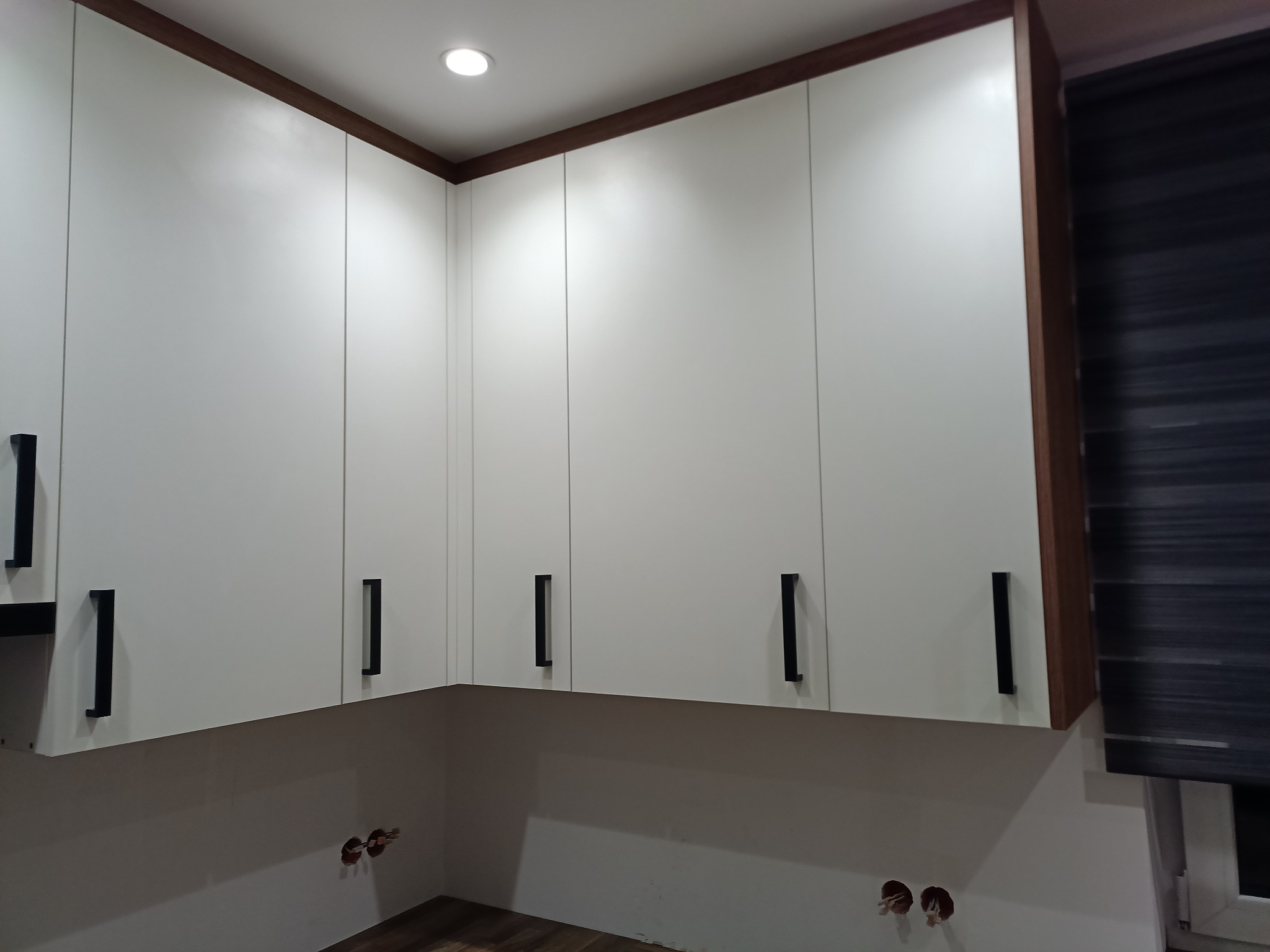 Białe szafki kuchenne wiszące z czarnymi, pionowymi uchwytami, brązowe wykończenie górnej krawędzi, widoczne gniazdka elektryczne bez osłon, podłoga imitująca drewno.