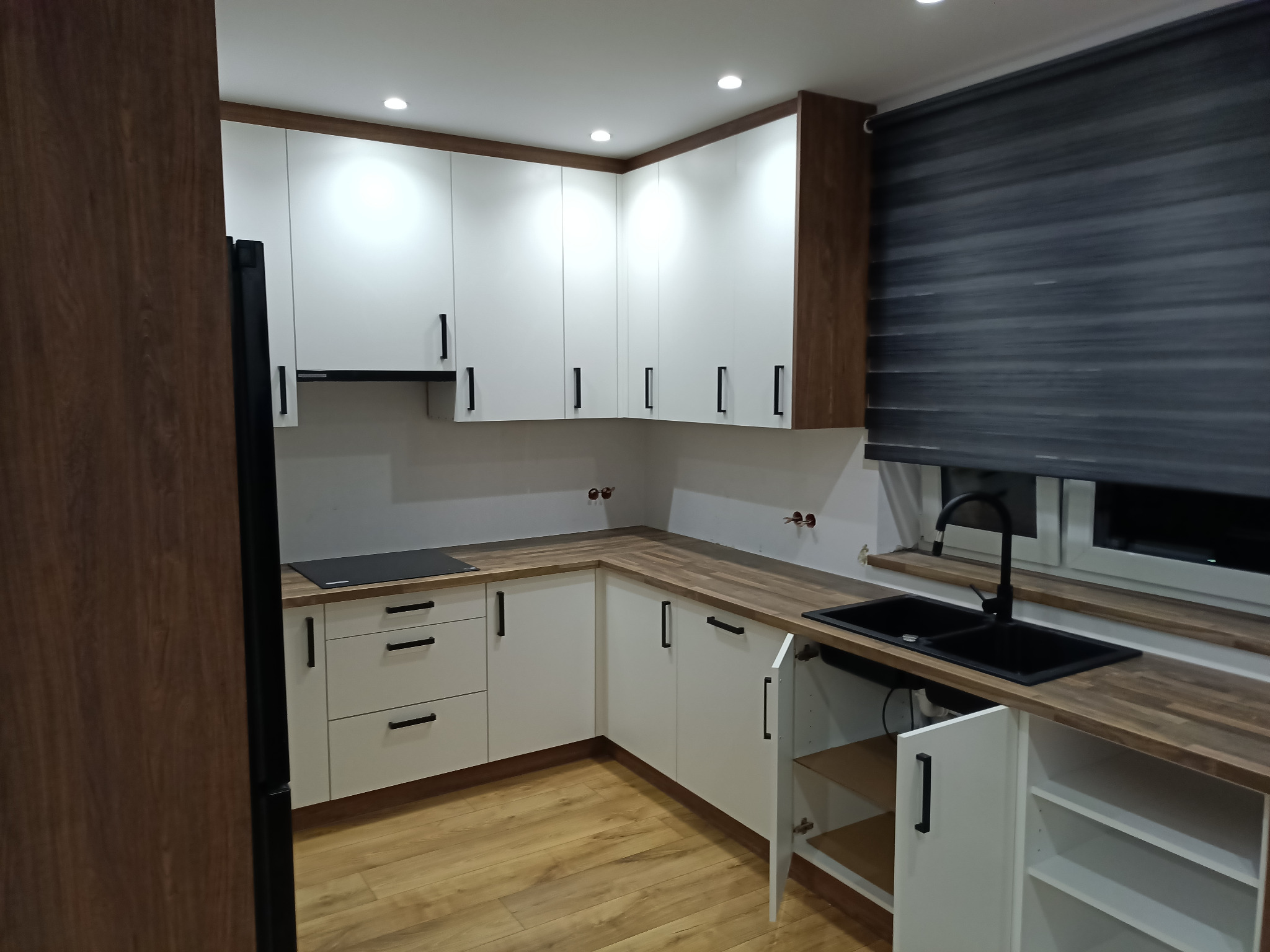 Nowoczesna kuchnia z białymi szafkami z czarnymi uchwytami, drewnianym blatem i czarnym zlewozmywakiem pod oknem z roletami, widoczne niedokończone podłączenia elektryczne.