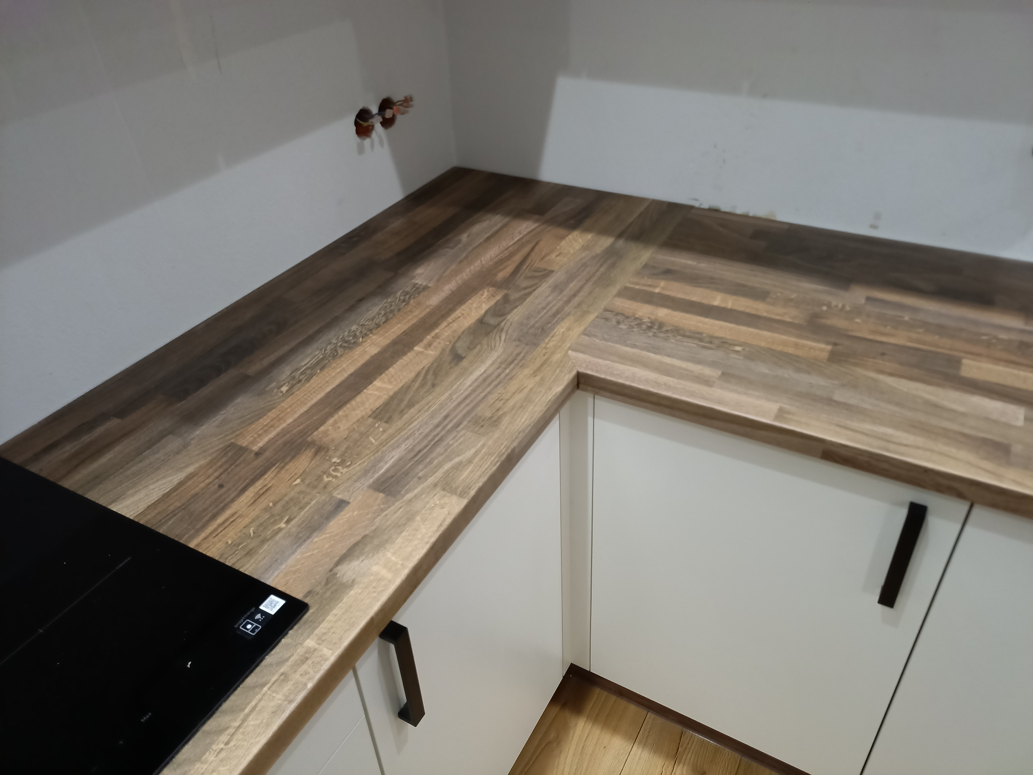 Fragment kuchni w trakcie wykańczania: drewniany blat z wbudowaną płytą indukcyjną na tle białych szafek z prostokątnymi, czarnymi uchwytami; widoczne niedokończone wykończenia ściany.