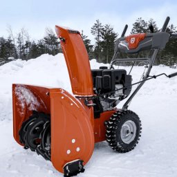 Mateusz Kura Dachy Płaskie - Pomarańczowy odśnieżacz spalinowy Husqvarna na śniegu, widoczne opony z bieżnikiem i logo producenta na uchwycie.