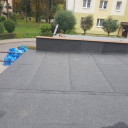 Mateusz Kura Dachy Płaskie - Widok z góry na płaski dach pokryty papą, z workami niebieskiej folii w rogu i budynkiem mieszkalnym w tle.