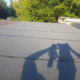 Mateusz Kura Dachy Płaskie - Płaski dach pokryty papą, widoczne łączenia i cień osoby robiącej zdjęcie, w tle drzewa i kominy wentylacyjne.