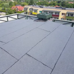 Szary, płaski dach pokryty papą termozgrzewalną, widoczny system wentylacyjny na dachu, w tle budynki mieszkalne i drzewa.