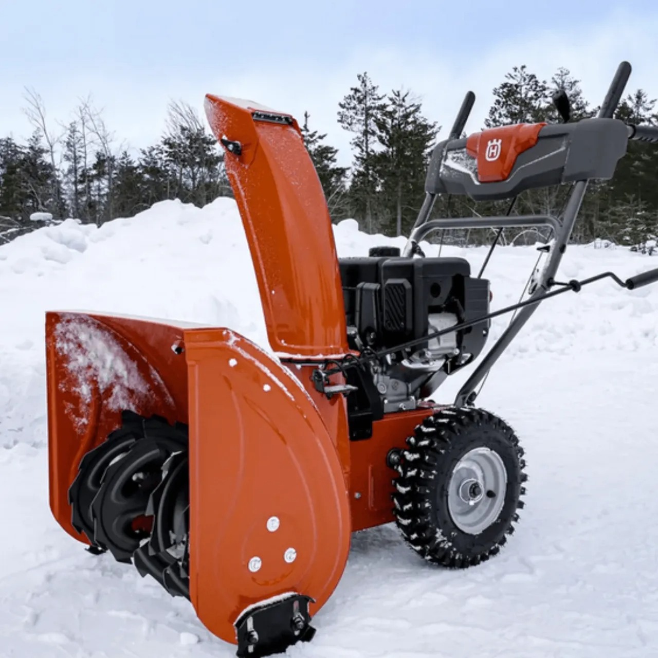 Pomarańczowy odśnieżacz spalinowy Husqvarna na śniegu, widoczne opony z bieżnikiem i logo producenta na uchwycie.