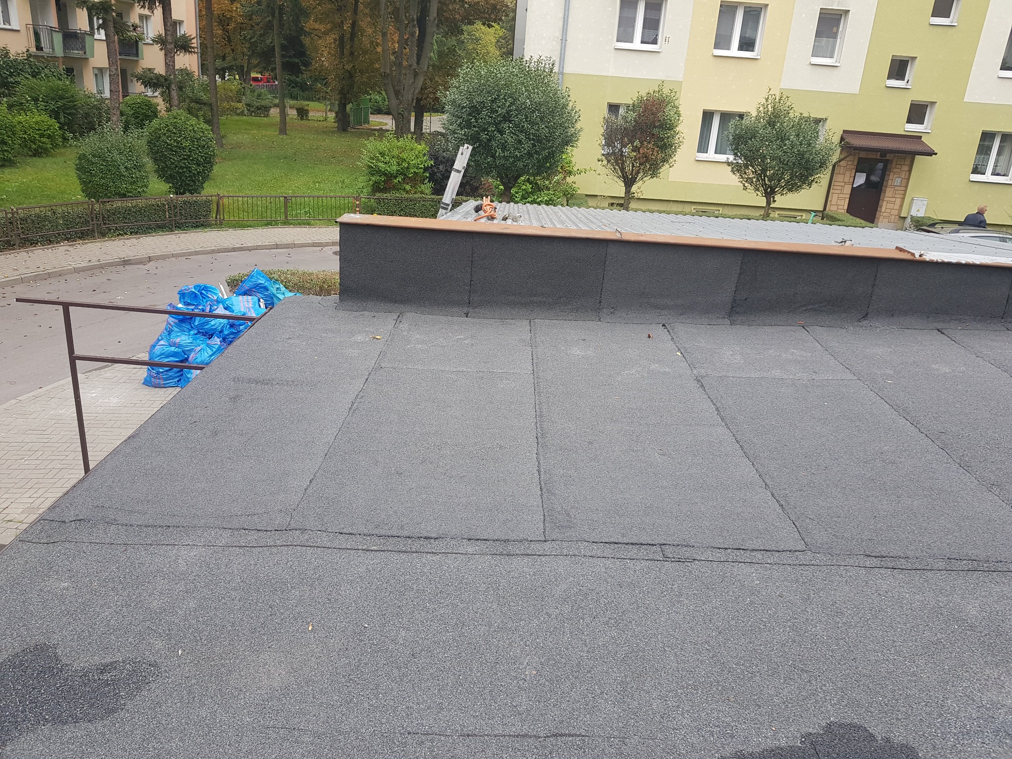 Widok z góry na płaski dach pokryty papą, z workami niebieskiej folii w rogu i budynkiem mieszkalnym w tle.