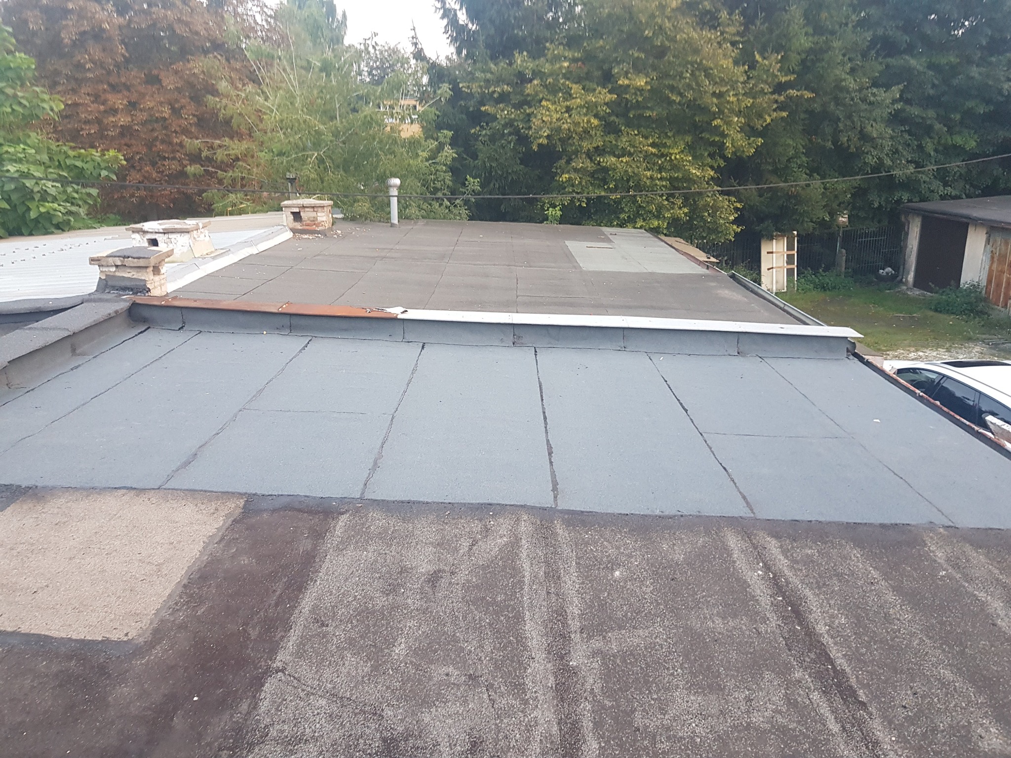 Płaski dach pokryty papą w różnych odcieniach szarości, z widocznymi kominami i elementami wentylacji, na tle zieleni drzew.