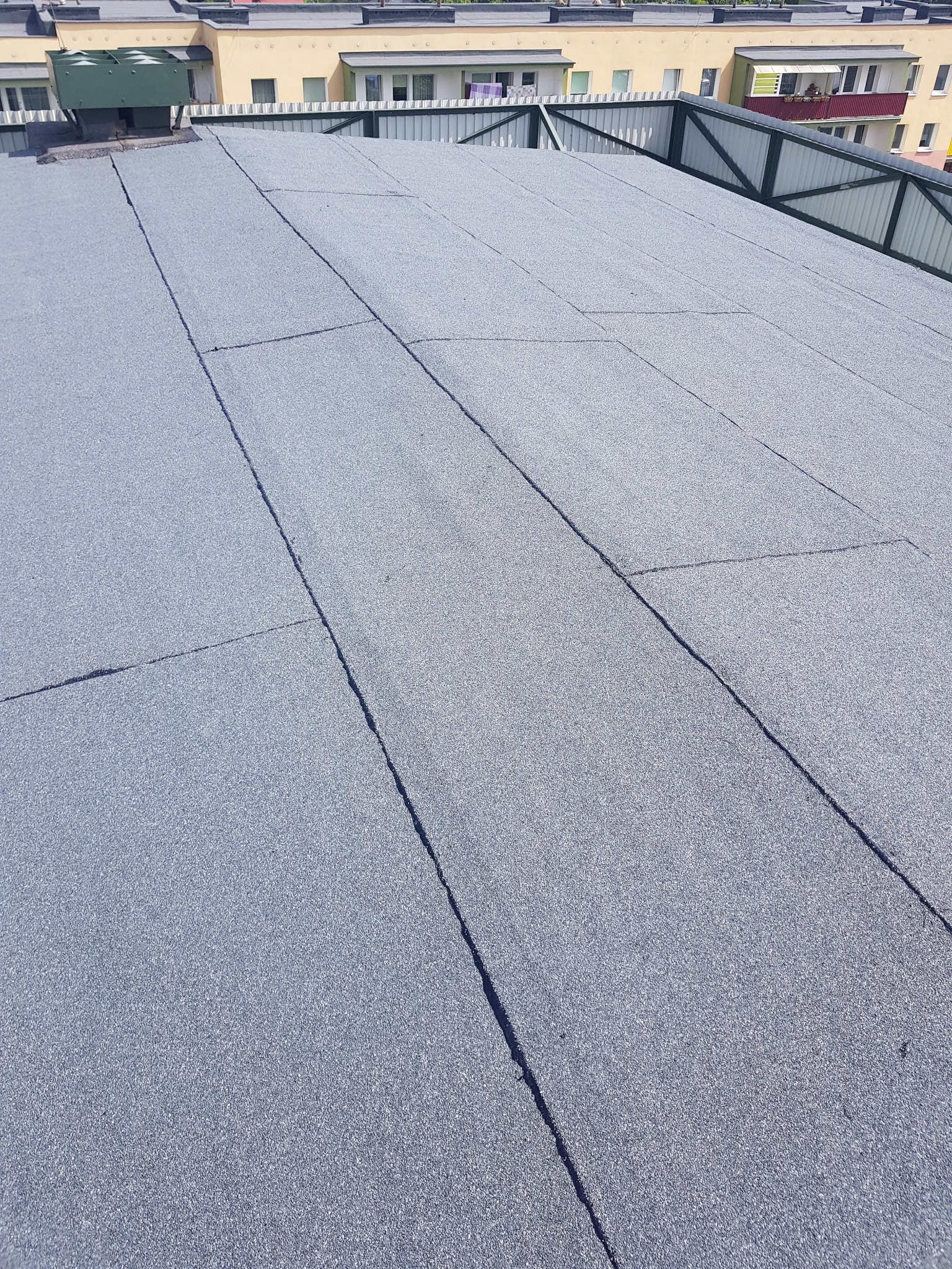 Płaski dach pokryty szarą papą termozgrzewalną, widoczne łączenia i budynki w tle.