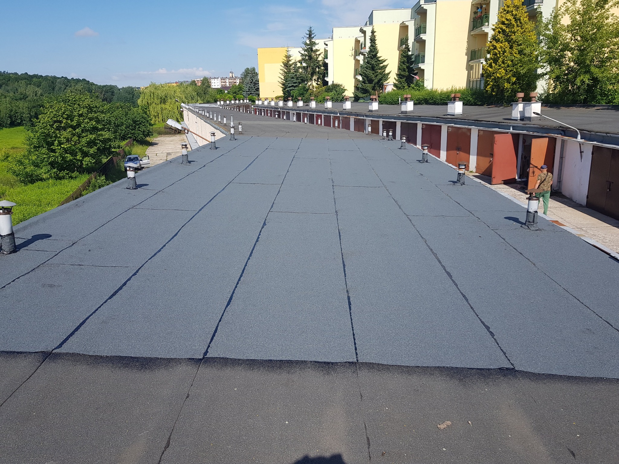 Nowe, szare pokrycie dachu płaskiego na budynku garażowym z kominkami wentylacyjnymi, na tle budynków mieszkalnych i zieleni.
