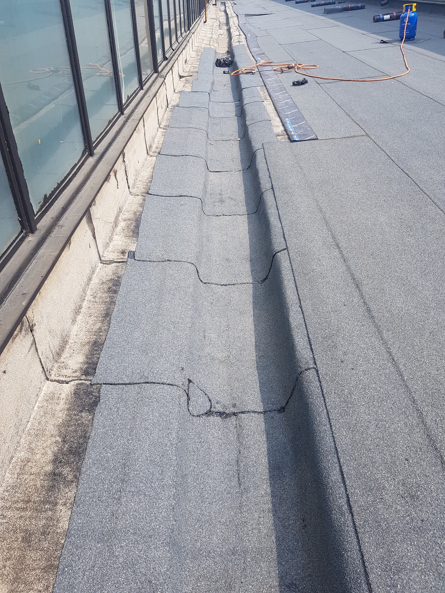 Bitumiczna papa na płaskim dachu budynku z widocznym systemem odprowadzania wody deszczowej i butlą gazową do zgrzewania papy.