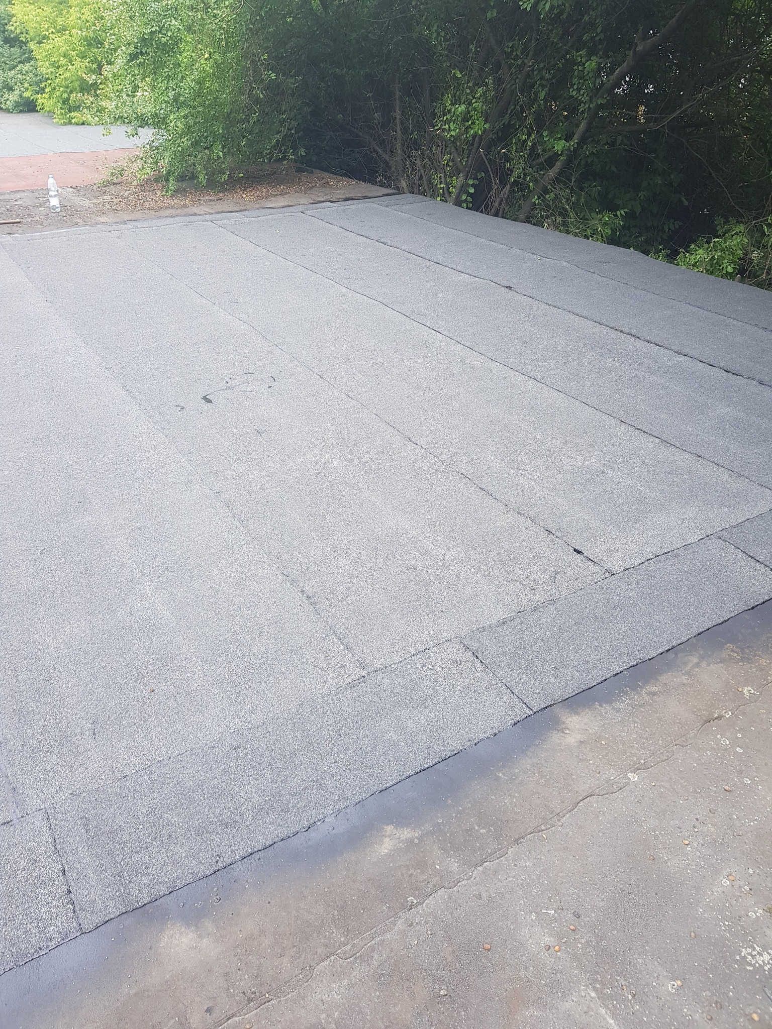 Płaski dach pokryty papą termozgrzewalną, widoczne łączenia i fragment roślinności w tle, butelka plastikowa w rogu dachu.