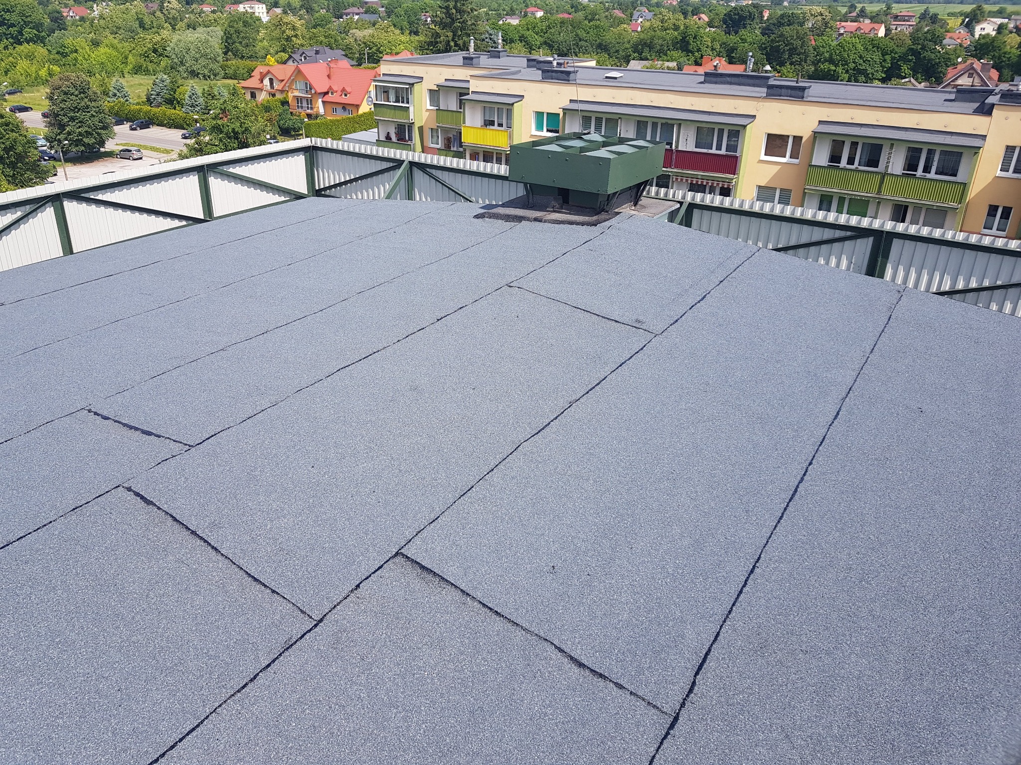 Szary, płaski dach pokryty papą termozgrzewalną, widoczny system wentylacyjny na dachu, w tle budynki mieszkalne i drzewa.