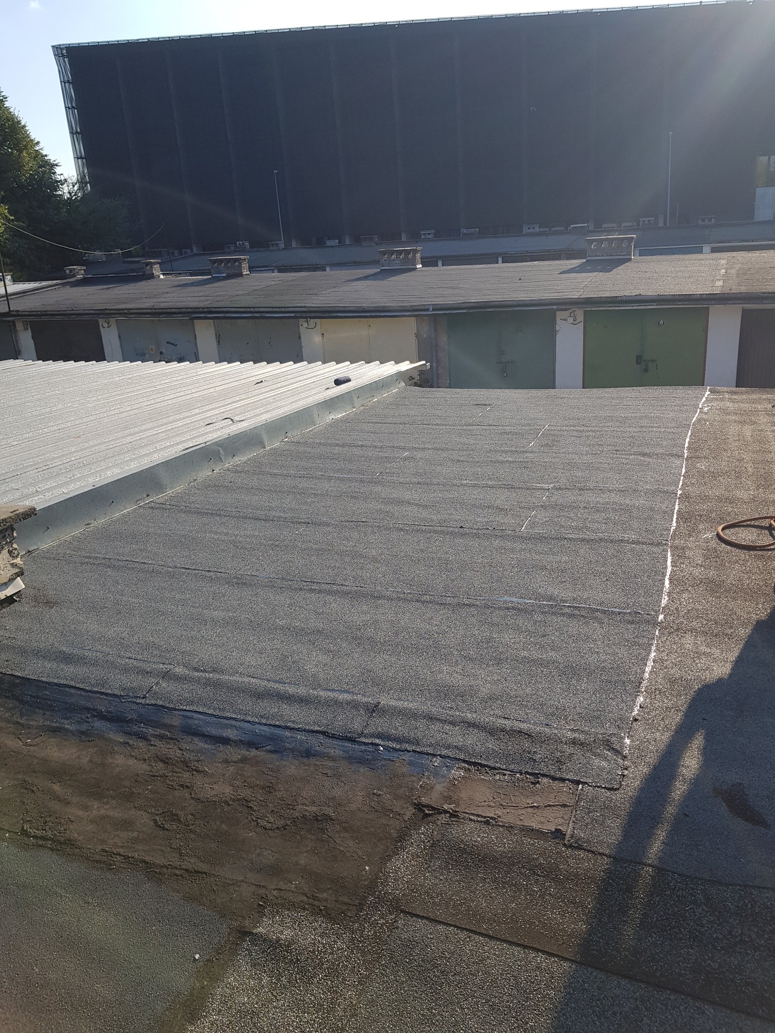 Widok z góry na płaski dach pokryty papą, z widocznymi garażami i budynkiem w tle.