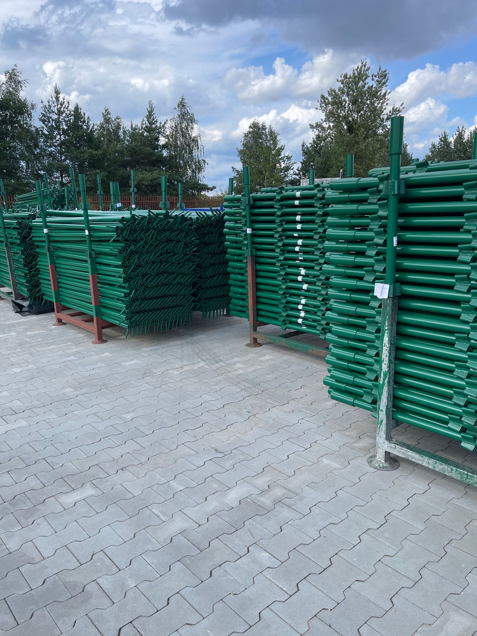 Stosy zielonych, metalowych słupków ogrodzeniowych ułożone na metalowych regałach na zewnątrz, na tle drzew i pochmurnego nieba.