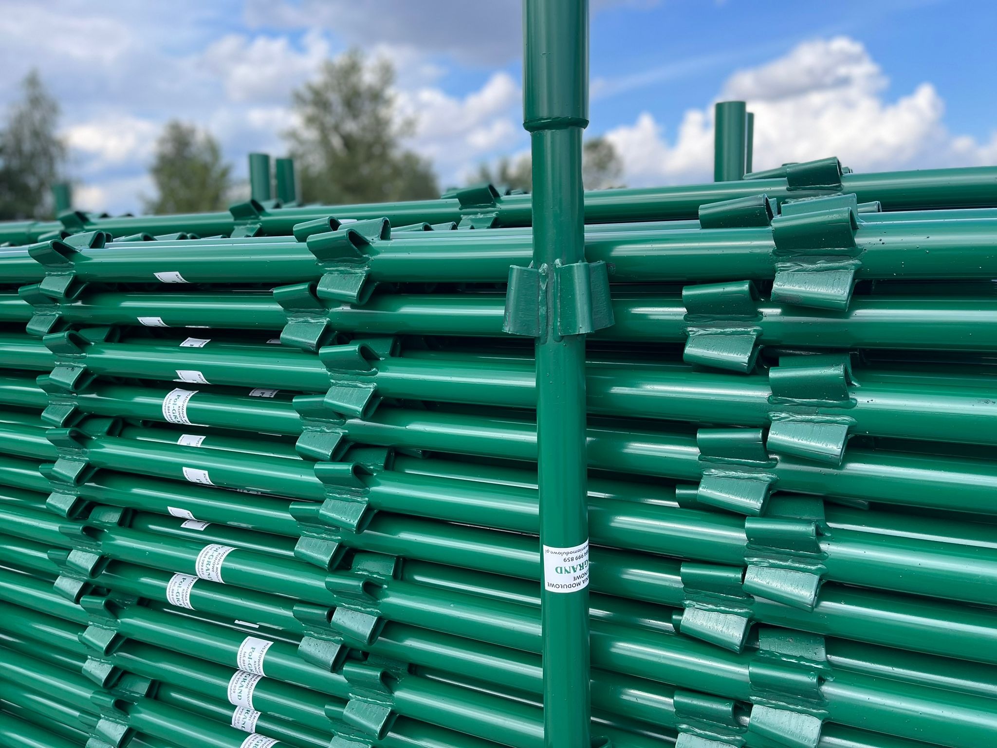 Stos zielonych, metalowych podpór budowlanych z widocznymi uchwytami mocującymi, ułożonych równolegle, z etykietami producenta, na tle błękitnego nieba z białymi obłokami i zielenią drzew.