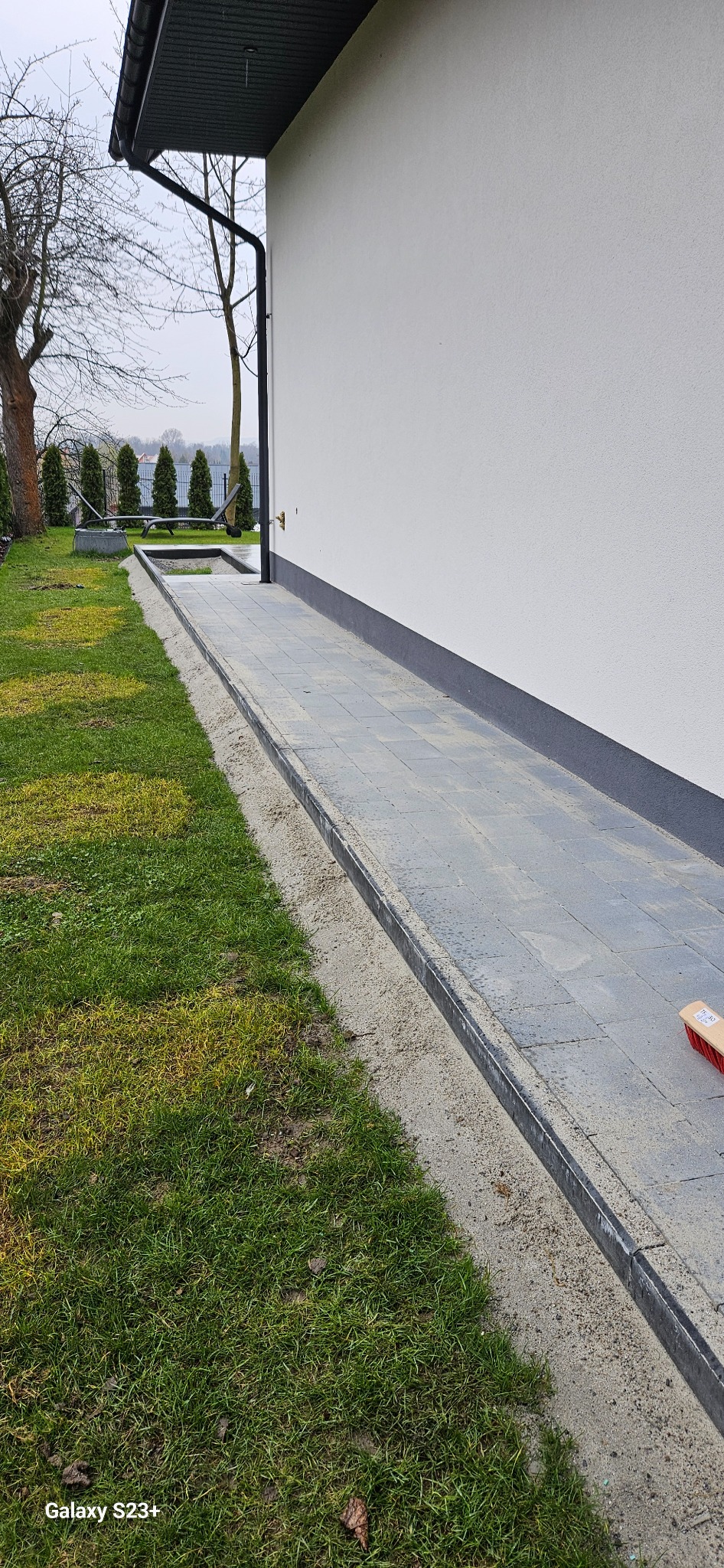 Ułożona ścieżka z szarych kostek brukowych przy białej ścianie budynku, obok trawnik, widoczny fragment szczotki do zamiatania.