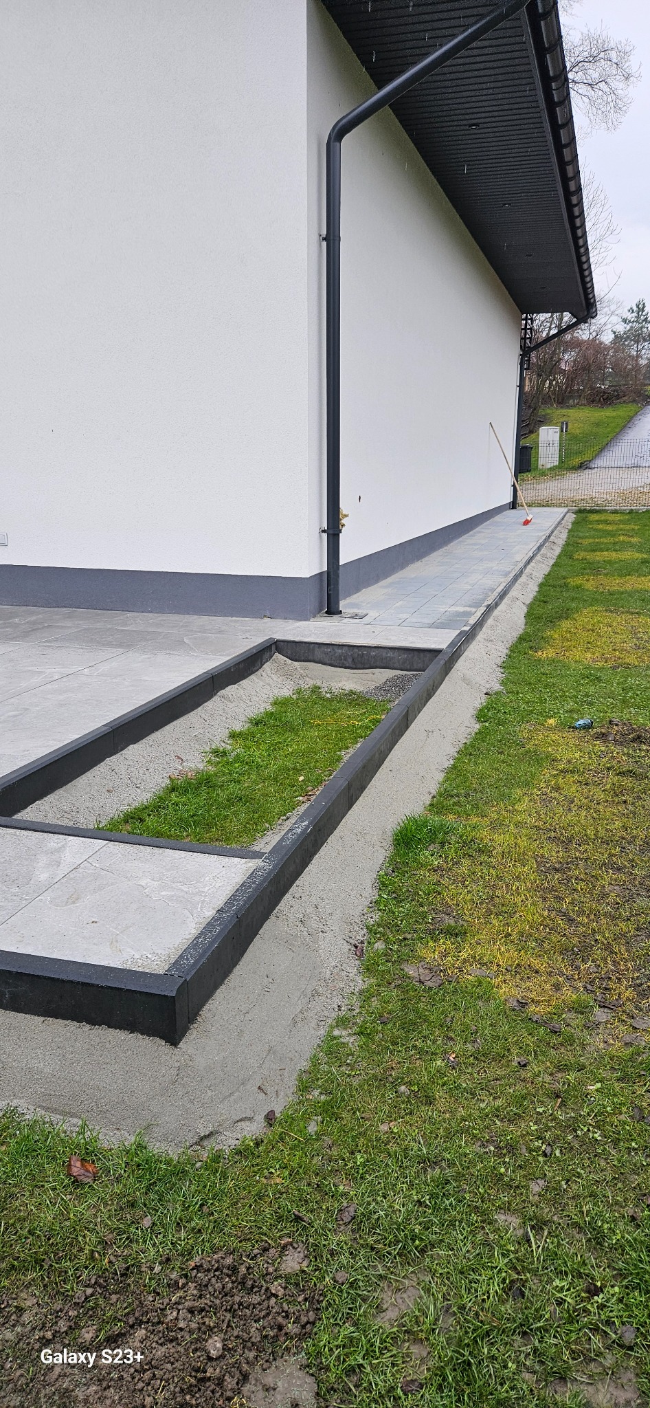 Wykończona ścieżka z kostki brukowej obok domu z fragmentem trawnika, w tle widoczna ściana budynku i rynna spustowa.