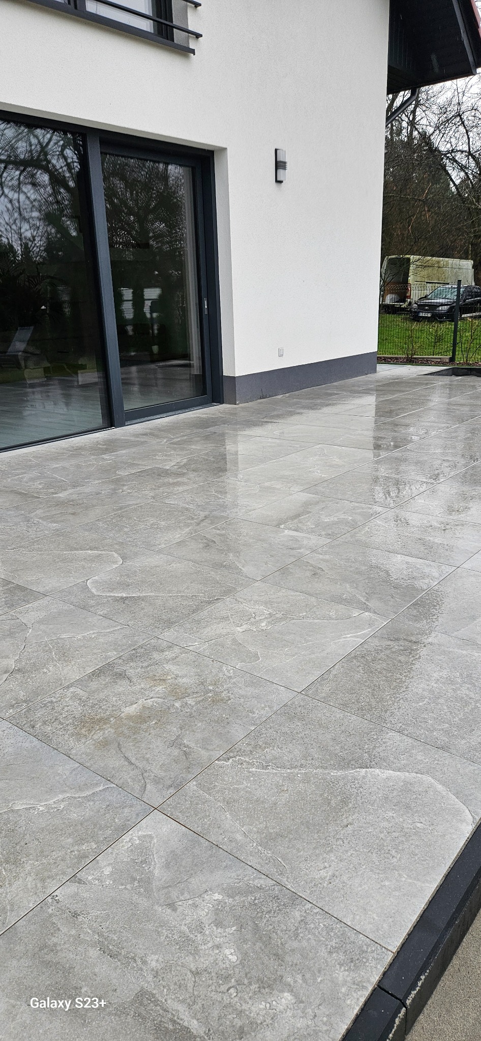 Ułożona, szara kostka brukowa imitująca kamień na tarasie w Bochni, widok z góry na mokrą nawierzchnię, zarys budynku z oknem przesuwnym w tle.