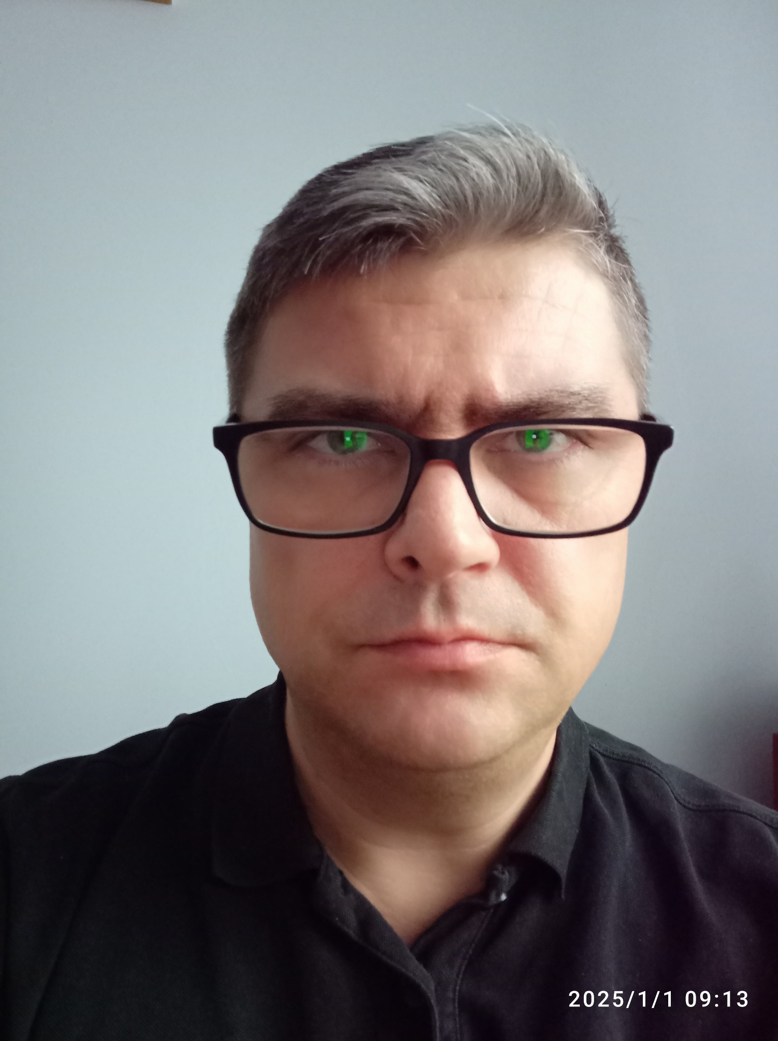 Portret mężczyzny w czarnej koszuli i okularach z czarnymi oprawkami, patrzącego wprost w obiektyw, z widocznym datownikiem w prawym dolnym rogu.