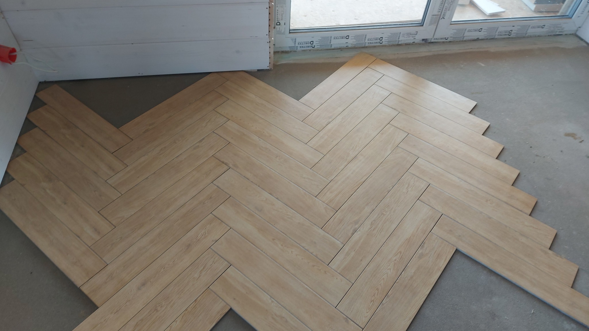Układanie jasnych, drewnopodobnych płytek w jodełkę na podłodze, widoczne niedokończone krawędzie i szara wylewka, okno w tle.