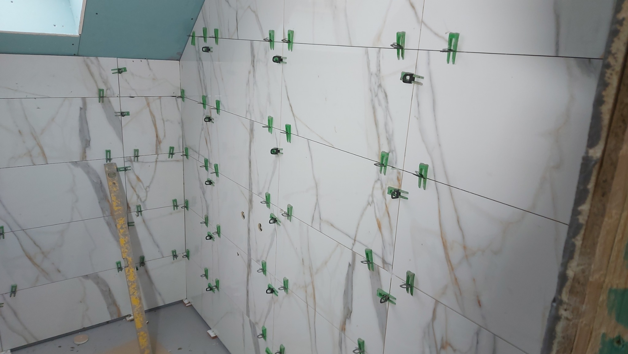Ściana w trakcie układania białych płytek imitujących marmur z widocznymi zielonymi klipsami poziomującymi i poziomicą przy ścianie.