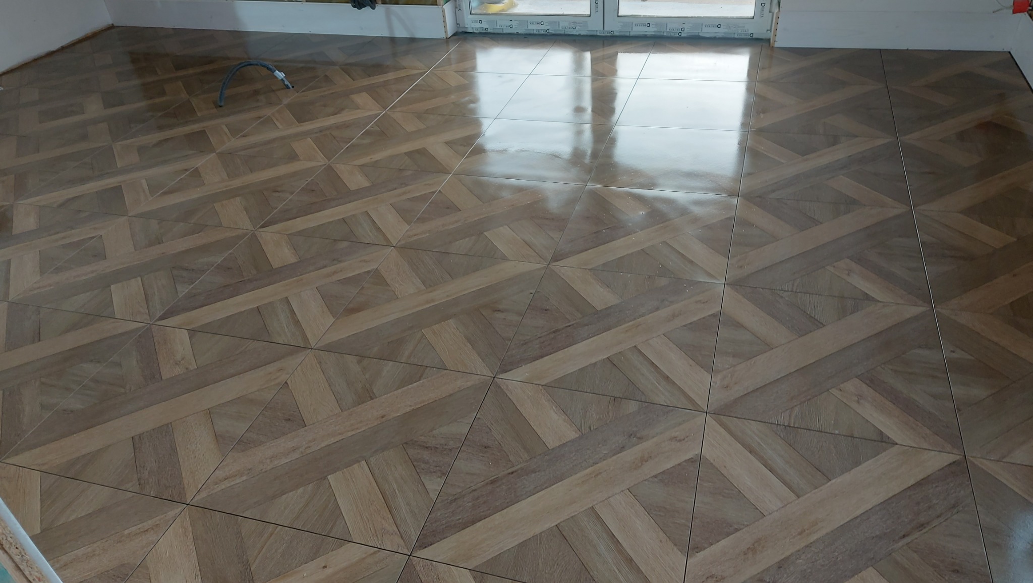 Ułożona podłoga z płytek imitujących drewno w jodełkę, widoczne łączenia i odbicia światła na powierzchni, kabel wystający ze ściany.