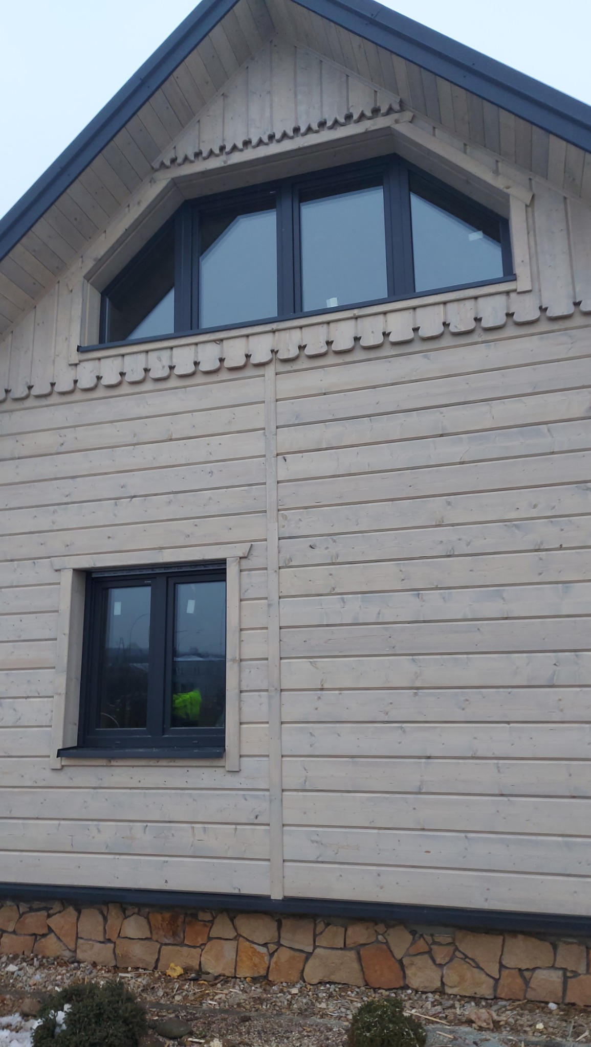 Fragment ściany zewnętrznej domu z jasnego drewna z dwoma oknami: trójkątnym na poddaszu i prostokątnym na niższej kondygnacji, wykończonej kamienną podmurówką.