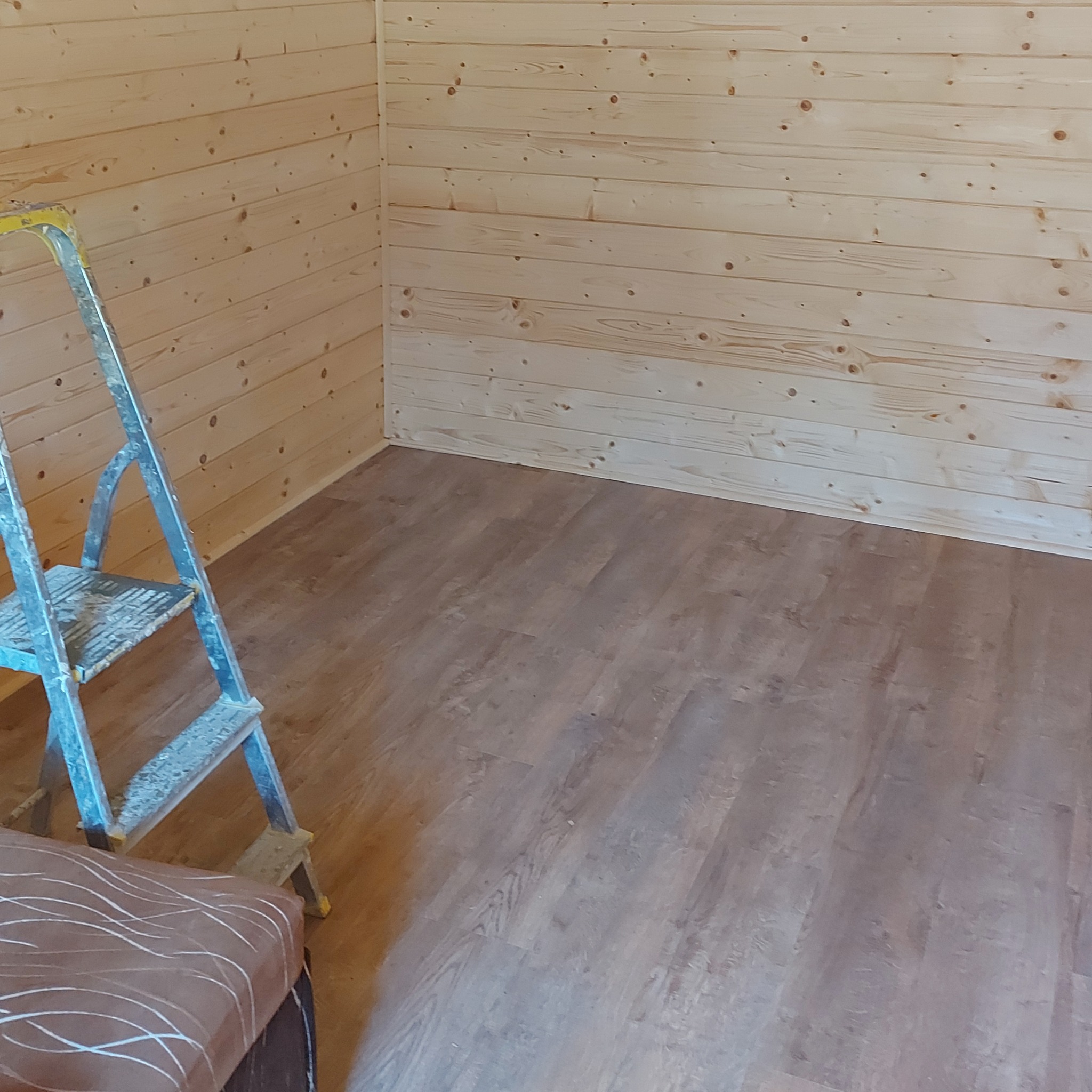 Wnętrze pomieszczenia z drewnianą boazerią i panelami podłogowymi, z widoczną aluminiową drabiną malarską w rogu.