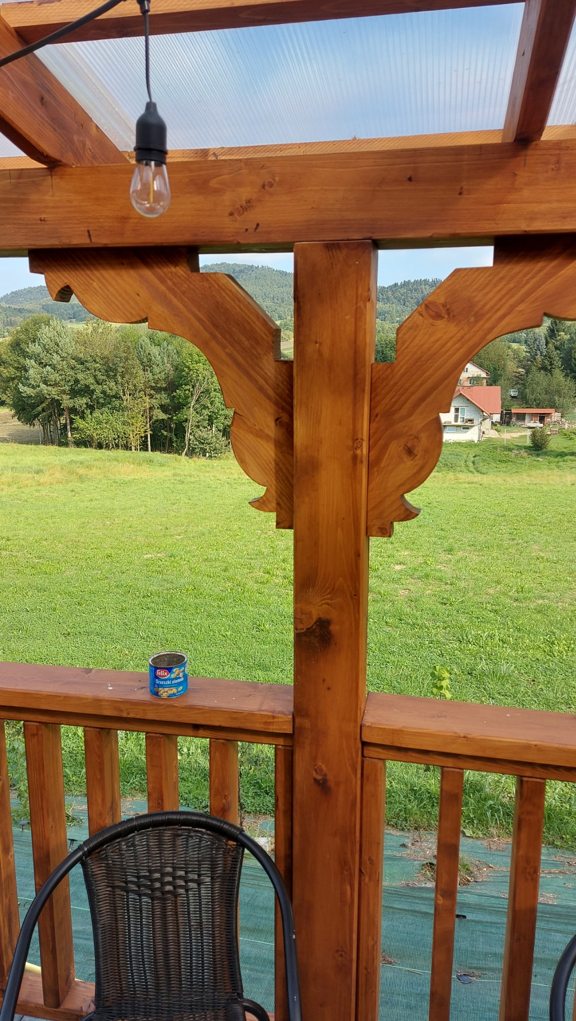 Drewniana konstrukcja tarasu z dekoracyjnymi wycięciami, widok na zielone wzgórza, puszka orzeszków na balustradzie i czarne krzesło w rogu kadru.