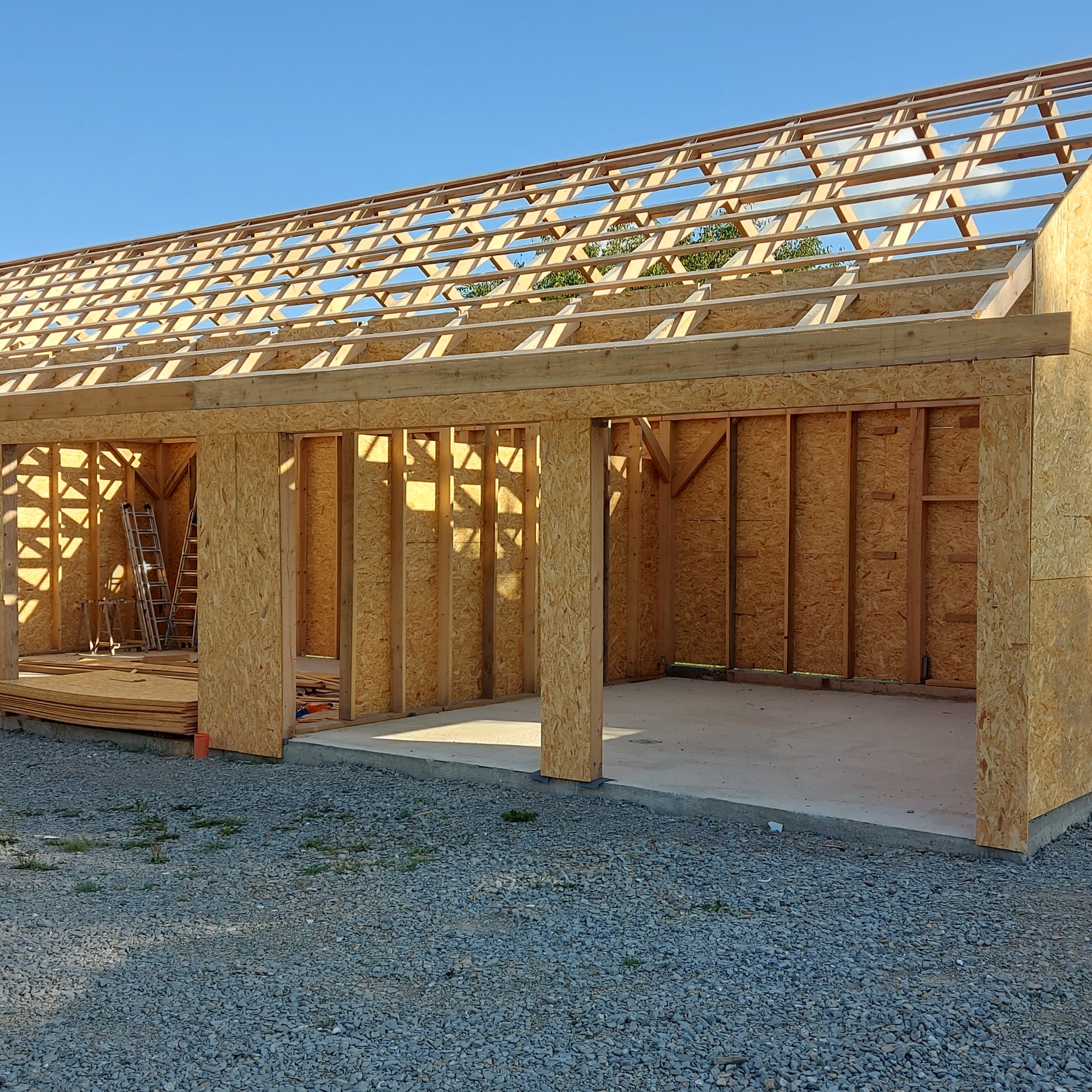 Konstrukcja szkieletowa budynku gospodarczego z widocznym drewnianym szkieletem ścian i dachu oraz betonową wylewką podłogi na żwirowym podłożu, w tle drabiny i płyty OSB.