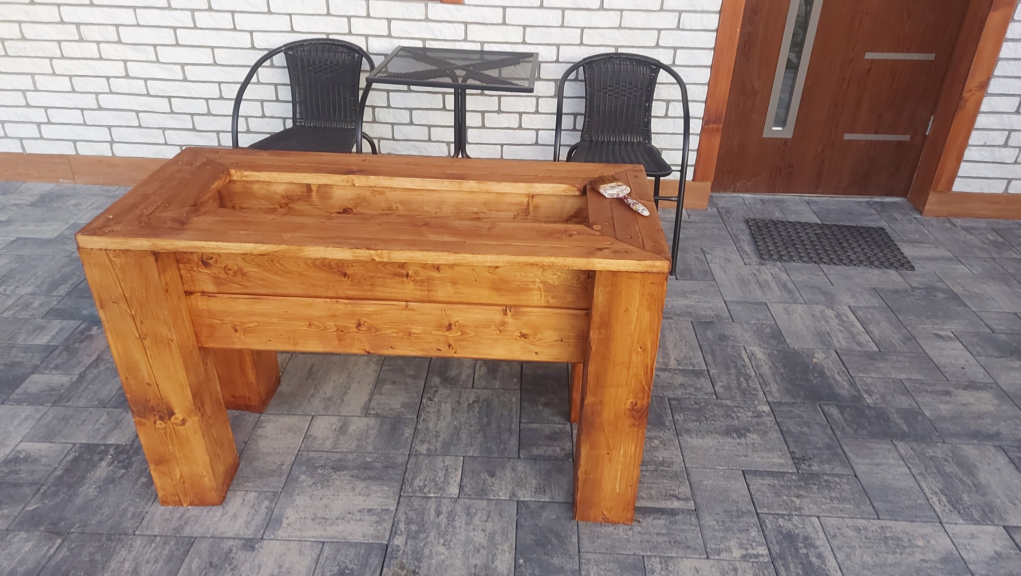 Drewniana donica na podwyższeniu, wykonana z bejcowanego drewna, z małą łopatką ogrodniczą na krawędzi, umieszczona na szarej kostce brukowej przed wejściem do domu z białej cegły.