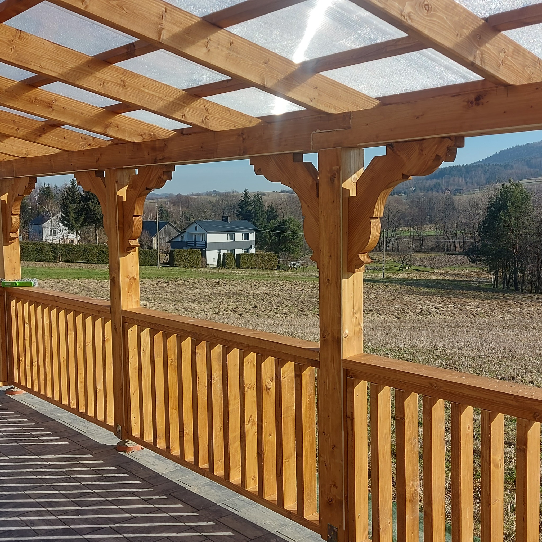 Drewniany taras z zadaszeniem z poliwęglanu, widok na krajobraz z domami w tle.