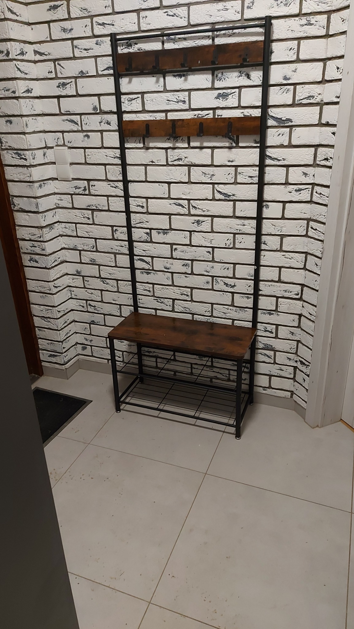 Metalowy wieszak z siedziskiem i półką na buty, ustawiony przy białej ścianie z imitacją cegły w przedpokoju.