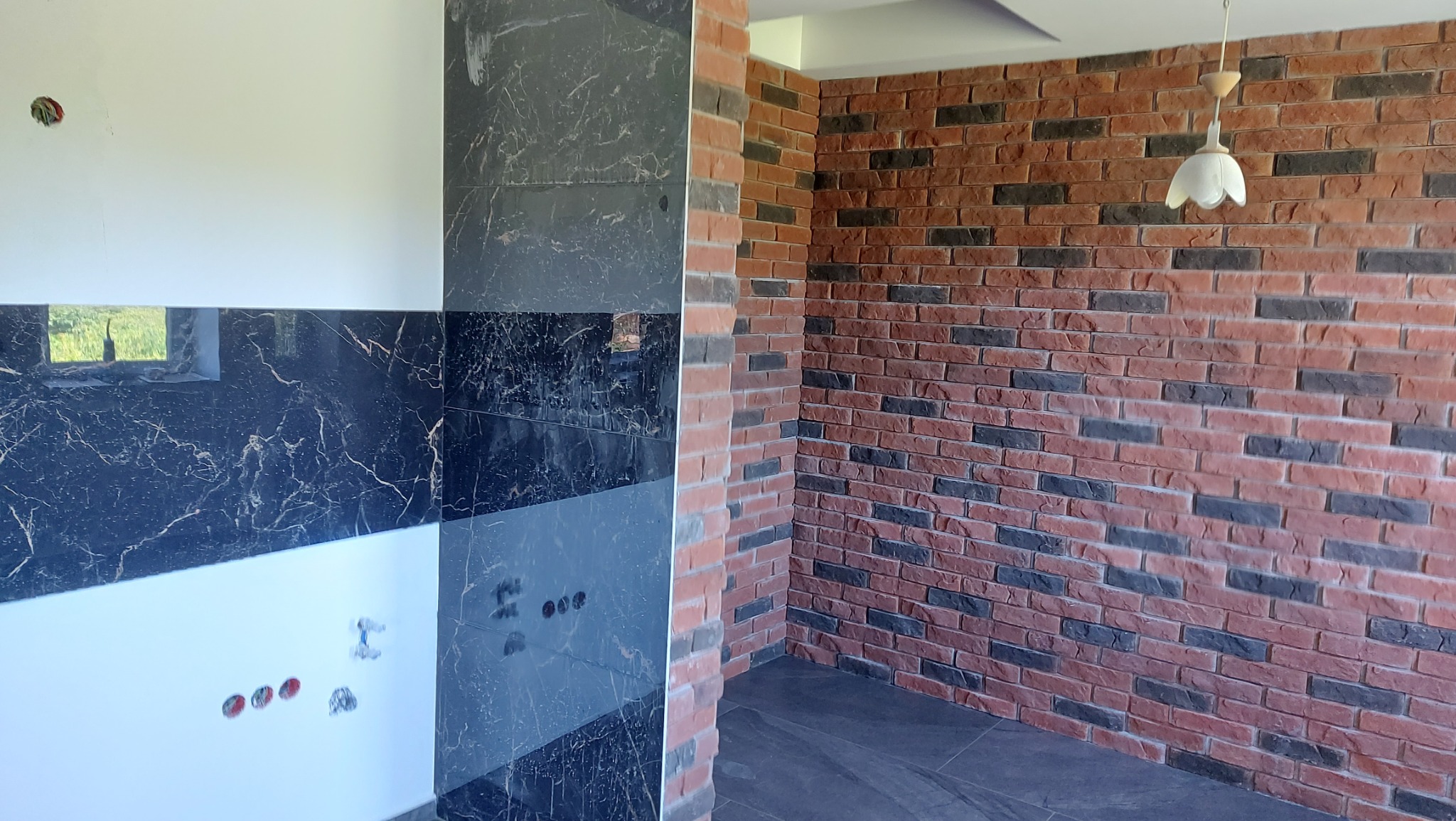 Fragment wnętrza z wykończoną ścianą z cegły dekoracyjnej w odcieniach czerwieni i szarości, sąsiadującą z ścianą wyłożoną czarno-białymi płytkami imitującymi marmur, widoczne gniazdka elektryczne...