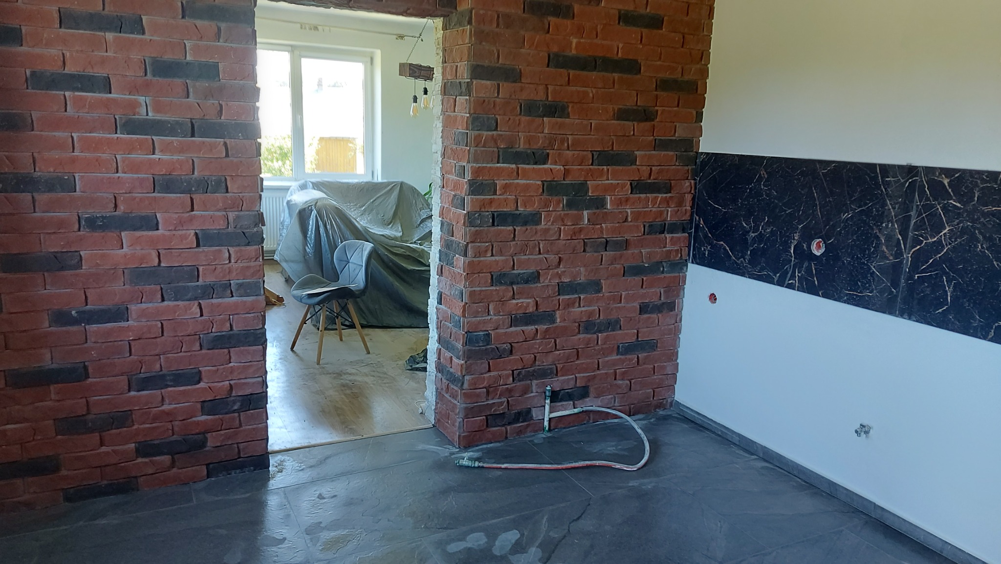 Wnętrze w Korczynie w trakcie prac wykończeniowych: fragment ściany z cegły klinkierowej, marmurowe panele ścienne i szara podłoga z płytek, widoczne instalacje.