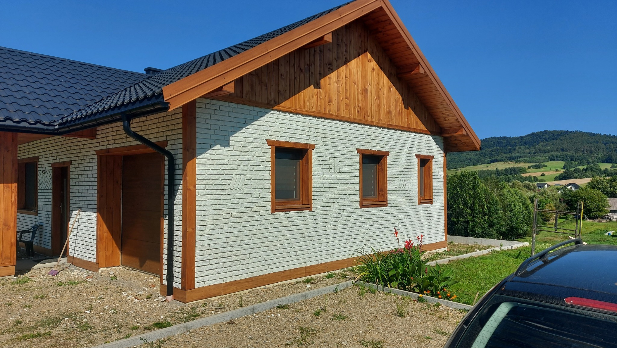 Dom z jasną elewacją imitującą cegłę, drewnianą stolarką okienną i wykończeniem dachu, na tle zielonych wzgórz i błękitnego nieba.